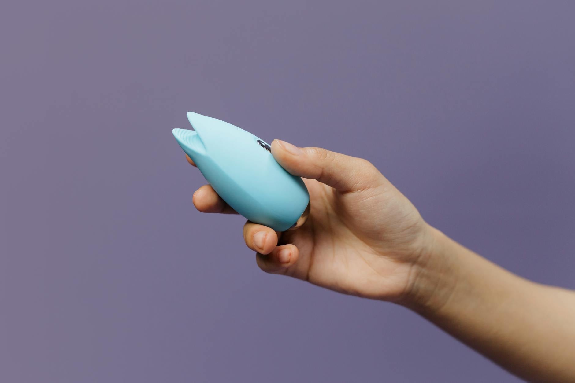 A hand holding a blue silicone suction vibrator against a soft purple background, symbolizing intimate self-care